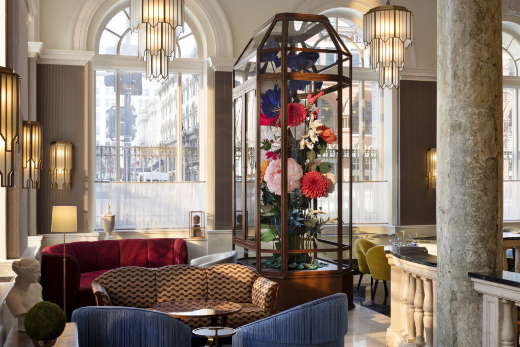 Lounge area at Cafe Riggs, situated between the dining room and the bar. Tall wood and glass display case in the centre holds an oversized paper flower arrangement. Curved velvet seating in red and brown patterned upholstery surrounds the case. Marble columns frame the space. Art deco wall sconces and pendant lights throughout.