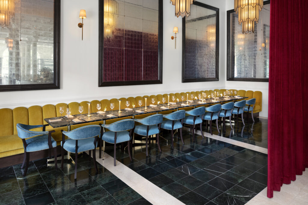 Private dining area at Cafe Riggs. Long marble-topped communal table set for up to twenty guests. Continuous mustard yellow banquette along the back wall with blue velvet chairs opposite. Three large antiqued mirrors in dark wood frames mounted above the banquette. Green marble floor with white marble inlay. Red velvet curtain on the right.