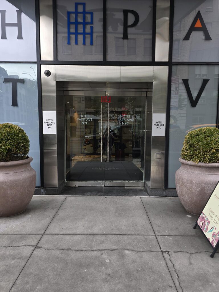 Street entrance at Hotel Park Ave NYC. Glass doors at pavement level with a level threshold, “Hotel Park Ave NYC” signage, planters either side.