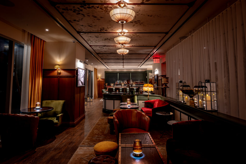 Golden Child lounge at Hotel Park Ave. Pendant fabric lights run the length of the room above seating. Red velvet armchairs and tan leather club chairs around low tables. Bar with green stools at the rear. Painted floral treatment on the ceiling continues over the bar. Wooden floor with patterned rug.