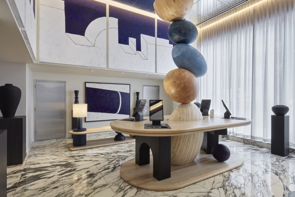 Reception area at Hotel Park Ave. Oval check-in desk in pale ash with a stacked wooden sculpture rising through the centre. Four screens and payment terminals set into the desk. Veined marble floor in black and white. Large abstract canvases on the wall behind. Floor-to-ceiling sheer curtain along the window line.