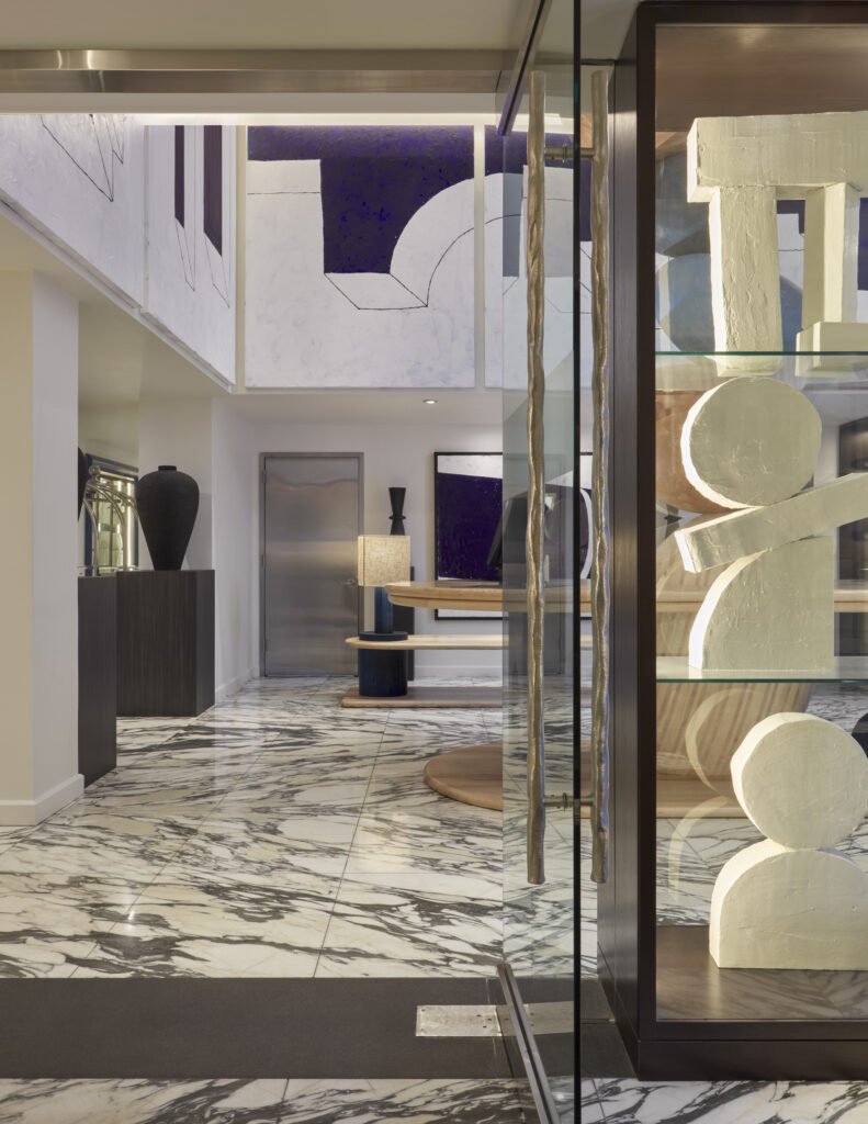 Lobby threshold at Hotel Park Ave looking through to reception. Glass-fronted display vitrine on the right holding cream sculptural objects. Bronze door handles. Marble floor continues into the reception area. Black plinth with a tall sculptural vessel on the left.
