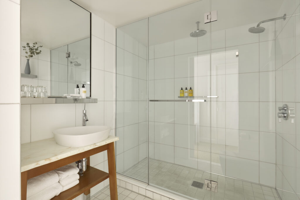 Studio Suite bathroom at Hotel Park Ave. Walk-in shower behind a fixed glass screen with two ceiling-mounted rain heads. Vessel basin on a marble-topped wooden vanity with folded towels on the shelf below. Wall-mounted shelf and mirror above the basin. White wall tile throughout.