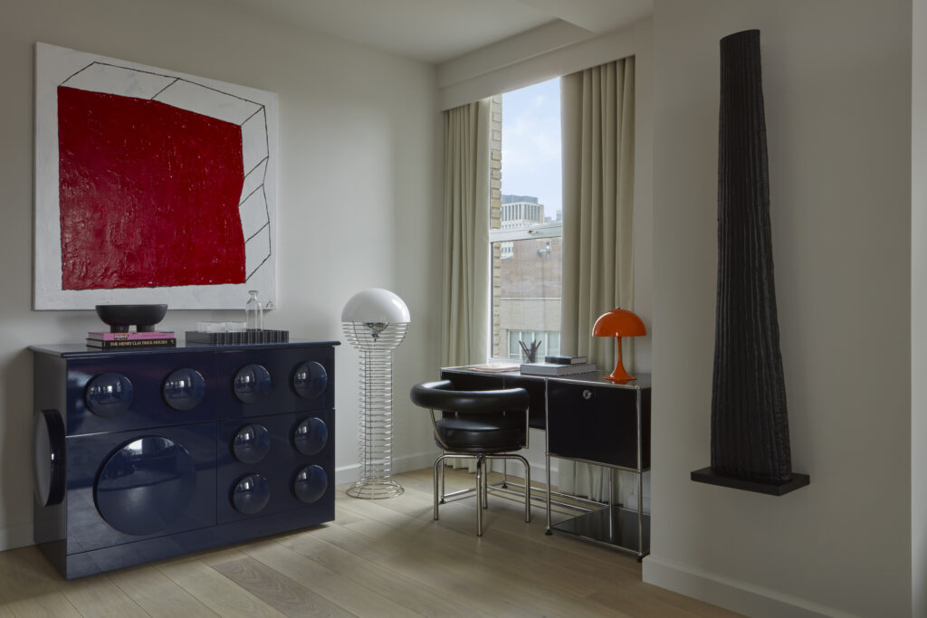 Studio Suite workspace at Hotel Park Ave. Dark desk and mounted shelving unit paired with a leather and chrome chair. Orange mushroom-shape table lamp on the desk. Large red abstract canvas on the wall above a dark blue lacquered cabinet with circular detailing. Wire-frame floor lamp beside the desk. Tall ribbed black sculpture mounted on the right.