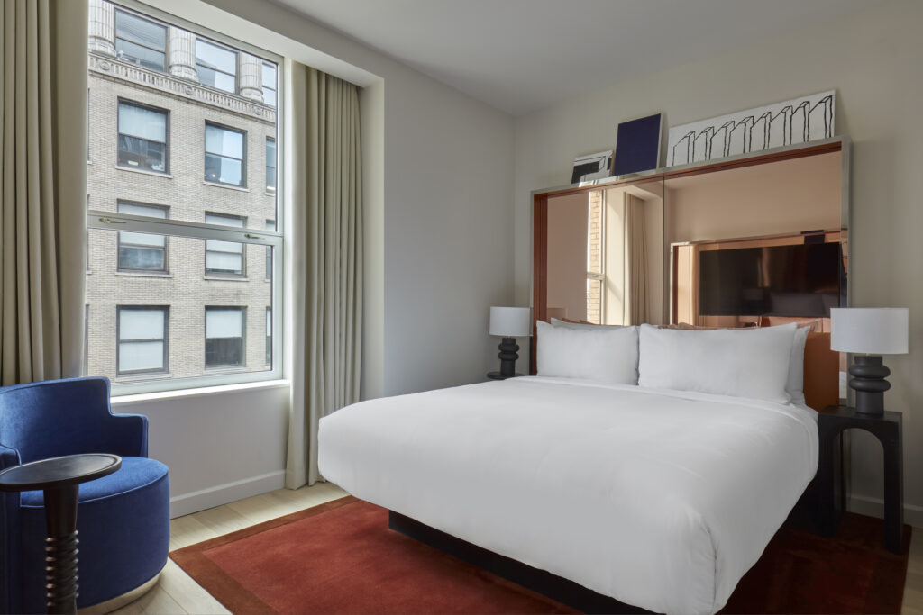 Studio Suite bedroom at Hotel Park Ave. King bed with white linens and an orange-red rug at the foot. Mirrored bronze headboard wall with a wall-mounted TV inset. Blue armchair with a small black side table beside a window onto an adjacent building. Sculptural black ceramic lamps either side of the bed. Wooden floor.