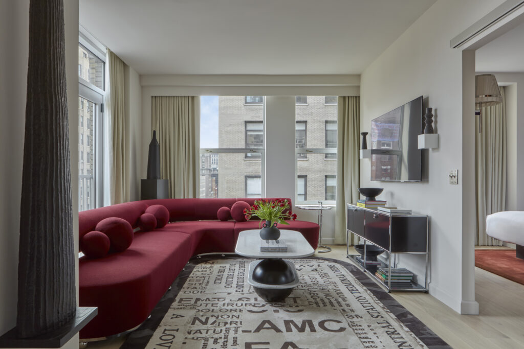 Studio Suite living area at Hotel Park Ave. Curved red sofa with spherical cushions. Round marble coffee table with a black vase of tulips. Wall-mounted TV opposite the sofa. View through to the bedroom past an open partition on the right. Tall ribbed black floor sculpture on the left. Newspaper-print rug across the floor.