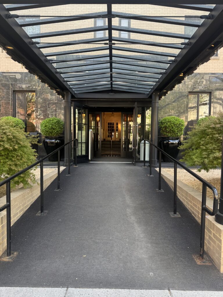 Entry ramp at Lyle Washington DC. A black non-slip surface ramp leads up from the pavement to the hotel doors. Black metal handrails run on both sides. A glass and steel canopy covers the full length of the ramp. Black ceramic planters with clipped shrubs flank the doors.