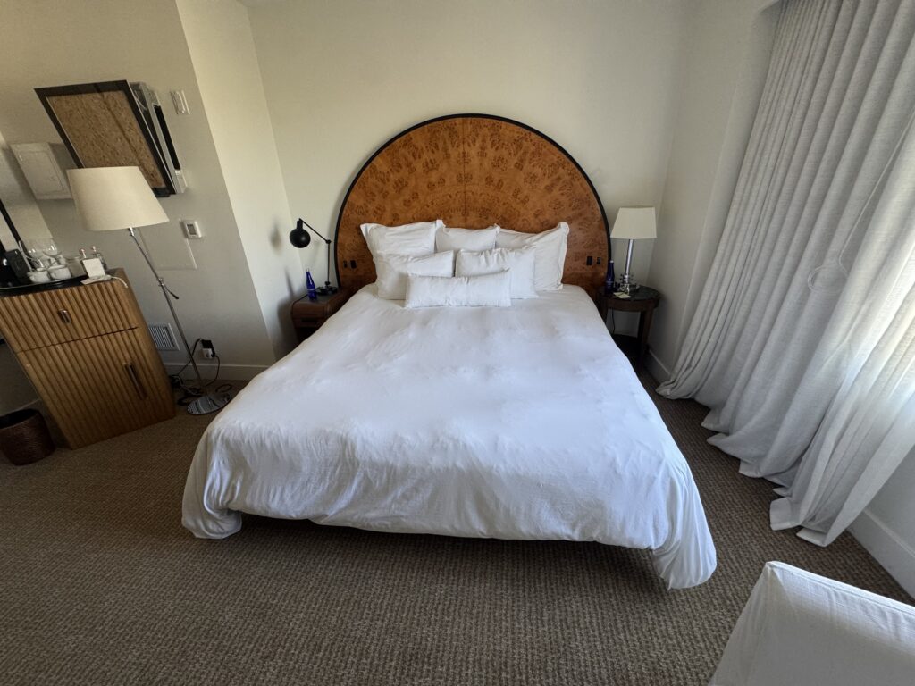 Accessible bedroom layout at Lyle Washington DC. A queen bed with arched burl-wood headboard sits centred on the wall. Carpet runs floor to wall. To the left, a ribbed wooden cabinet sits beside a swing-arm floor lamp. Clear floor space runs along the side and foot of the bed.