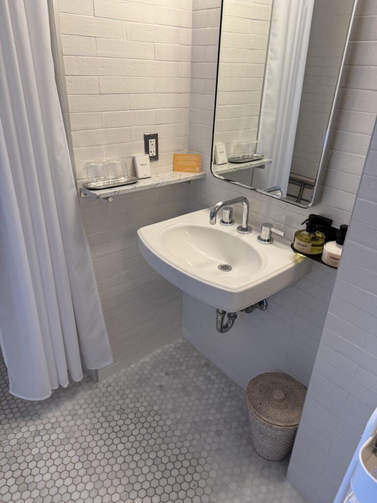 Accessible bathroom sink at Lyle Washington DC. Wall-mounted ceramic sink with exposed chrome plumbing below for wheelchair clearance. A marble shelf with vanity glassware sits above the sink. Chrome single-lever taps are mounted on the sink rim. Hex-pattern marble mosaic floor runs throughout.