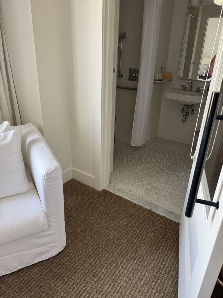View from the accessible bedroom into the bathroom at Lyle Washington DC. The bathroom doorway has a low marble threshold strip running across the entry. Beyond the threshold, the roll-in shower with curtain is visible to the left. The wall-mounted accessible sink is visible to the right. Carpet runs up to the threshold; the bathroom floor is hex-pattern marble mosaic.