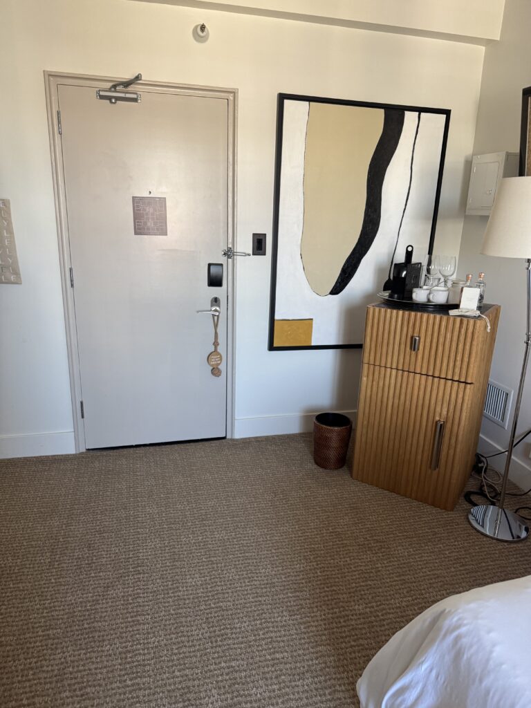 Bedroom entry door at Lyle Washington DC viewed from inside the accessible bedroom. The door is solid with a lever handle and a door closer mounted at the top. An electronic keypad lock sits below the handle. An emergency evacuation plan is mounted at eye level. To the right, the room’s minibar in ribbed wood holds glassware and a coffee setup.