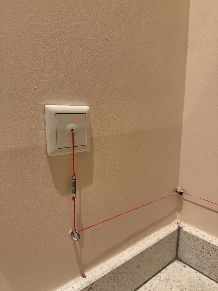 Emergency alarm activation point in the accessible public toilet at Kimpton de Witt. White wall plate with red indicator light and a red pull cord with a triangular hand grip, cord routed to floor level and to a corner anchor.