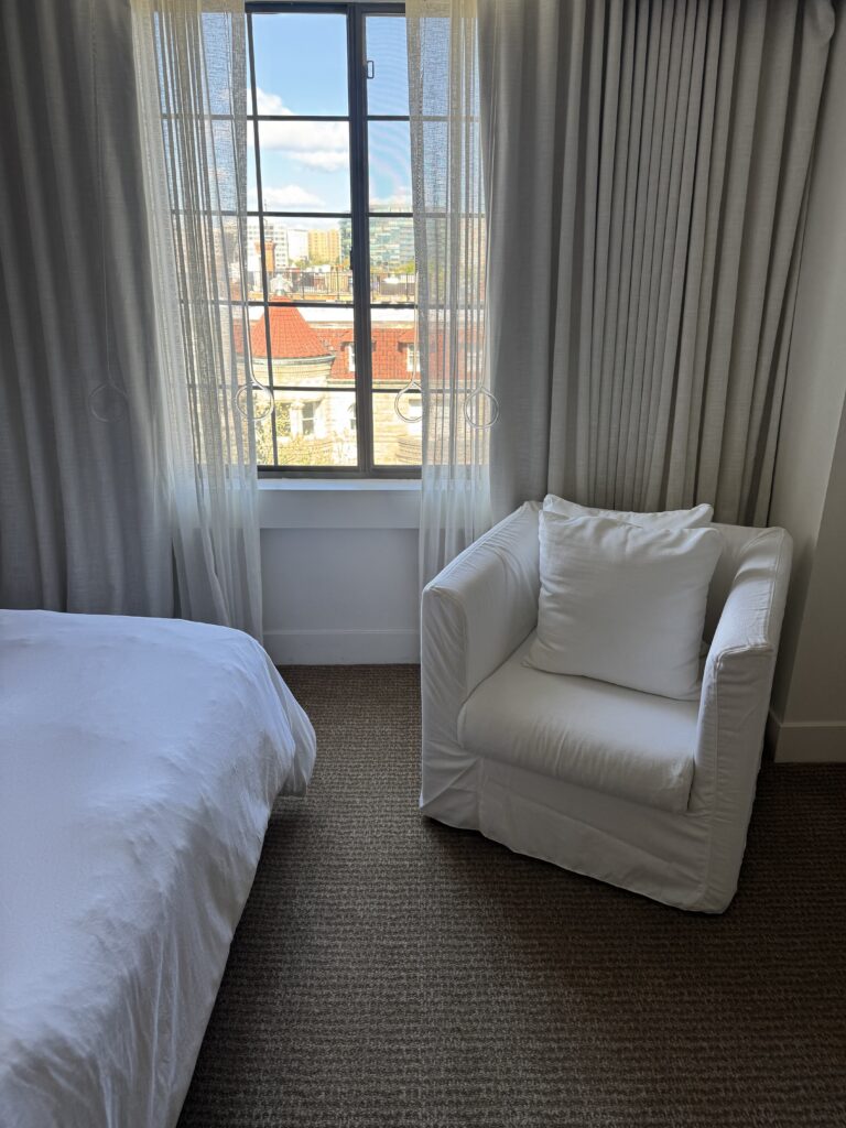 Bedroom window in the accessible room at Lyle Washington DC. Black-framed casement window with sheer and heavier linen curtains layered. A white slipcovered armchair with cushion sits in the corner. View shows Washington DC rooftops including a red-tiled turret. Carpet runs floor to wall.