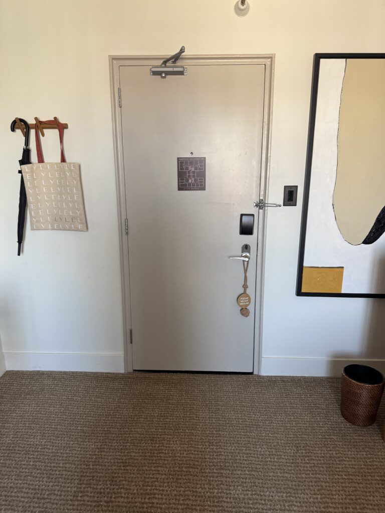 Bedroom entry door at Lyle Washington DC, wider view. Door closer mounted at the top. To the left of the door, wall-mounted coat hooks hold a black umbrella and a Lyle-branded canvas tote. An emergency evacuation plan and electronic keypad lock are mounted on the door.