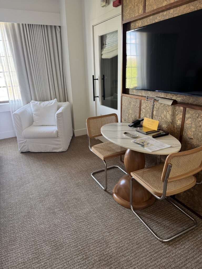 Workspace and seating area in the accessible bedroom at Lyle Washington DC. Two cane Cesca chairs with chrome legs sit at a round marble-topped pedestal table. A wall-mounted television is set into cork-clad cabinetry. A white slipcovered armchair sits to the left beneath full-length curtains. A swing-arm task lamp is mounted on the cabinet beside the television.