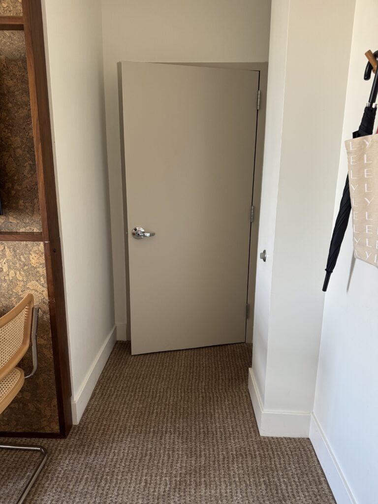 Accessible bathroom door from the bedroom side at Lyle Washington DC. A solid hinged door in pale beige with a chrome lever handle. The doorway has a step-free threshold transitioning from carpet to bathroom flooring. Cork-clad cabinetry of the television wall is visible to the left.
