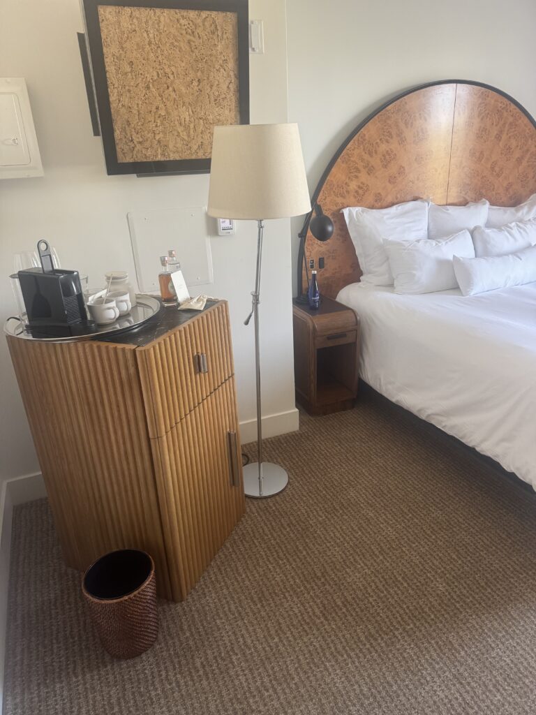 Bedside area in the accessible bedroom at Lyle Washington DC. A ribbed wooden minibar cabinet with a black marble top holds a coffee maker and glassware. A swing-arm reading lamp in black metal is mounted on the headboard. A wall-mounted thermostat sits on the wall to the right of the headboard. A bedside table in dark wood holds bottled water beneath the lamp.