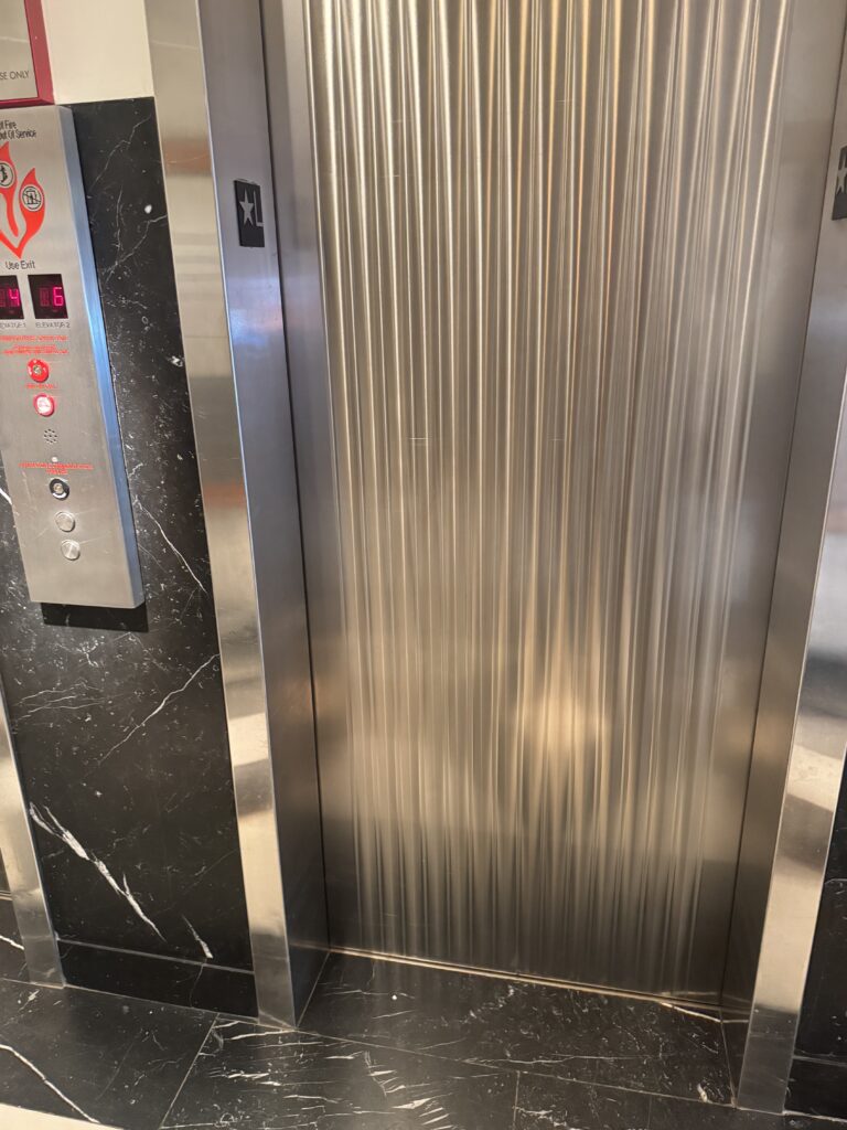 Lift exterior at Lyle Washington DC. Brushed stainless steel reeded lift doors marked with star and L for lobby, set in a black marble surround. Floor indicator display shows the current floor. Emergency fire recall buttons and elevator communication panel are mounted below the indicator.