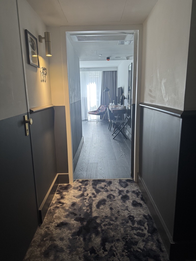 Corridor approach to a guest room at Kimpton de Witt. Dark dado-panelled walls, patterned corridor carpet meeting the room’s wood floor at a level threshold. Door open showing the room beyond.