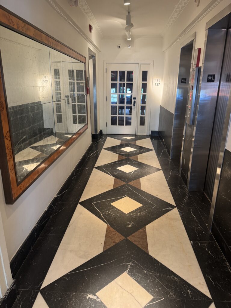 Lift bank and corridor at Lyle Washington DC. Two passenger lifts with brushed steel doors marked with star and L for lobby. The corridor floor is geometric marble inlay in dark and pale tones with deeper accent stones. A burl-wood-framed mirror hangs on the left wall. Internal glazed doors lead to the public areas at the end of the corridor.