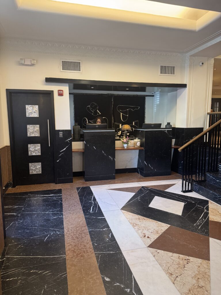 Reception desk at Lyle Washington DC. Two black marble check-in podiums with marble surfaces at standing height. Behind the desks, a tapestry depicts abstract figures in black and white. A black panelled door with art deco silver relief medallions sits to the left. Floor is marble inlay.