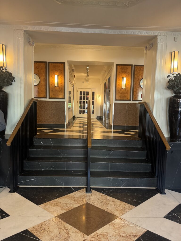 Internal lobby steps at Lyle Washington DC. Four marble steps between lobby levels with black metal balustrades and oak handrails. A central pillar with handrail divides the stair into two lanes. Burl-wood wall panels frame the steps with integrated wall sconces.
