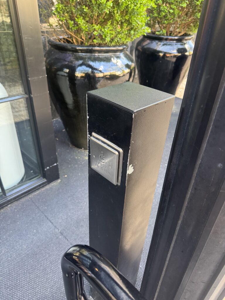 Push-to-open accessible entry button at Lyle Washington DC. A black metal column at the hotel entrance houses a stainless steel push pad with wheelchair symbol. The column sits beside the glass entry doors. A black ceramic planter with clipped boxwood sits behind.