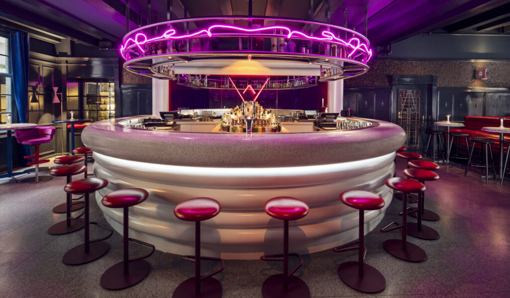 Super Lyan cocktail bar at Kimpton de Witt with circular central bar and red leather stools beneath a suspended magenta neon ring, set against dark wood panelled walls.