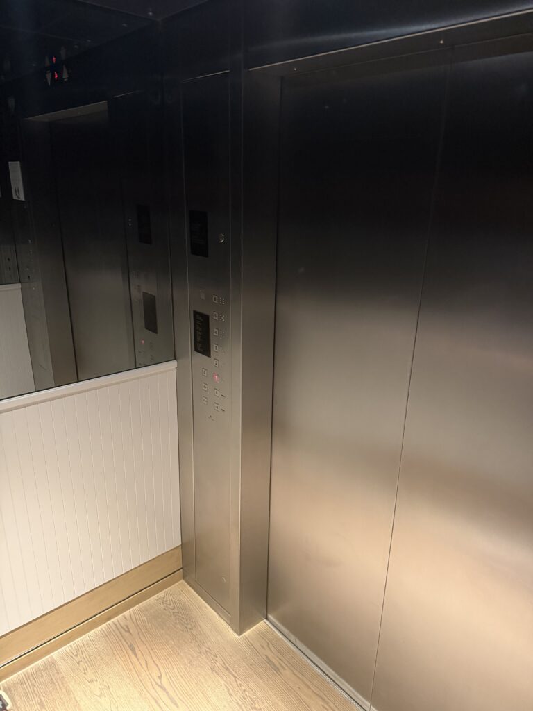 Lift cab interior showing control panel beside mirrored back wall, wood floor.