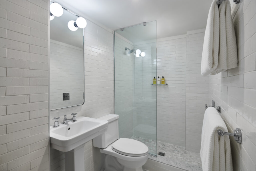 Bathroom at Lyle Washington DC. Walls are clad in white tile with a brick-effect joint pattern. A frameless walk-in shower has a fixed showerhead and glass toiletry shelf. The shower floor is hex-pattern marble mosaic. A traditional pedestal sink with cross-handle taps sits beside the toilet. Twin chrome towel rails are mounted on the right-hand wall.