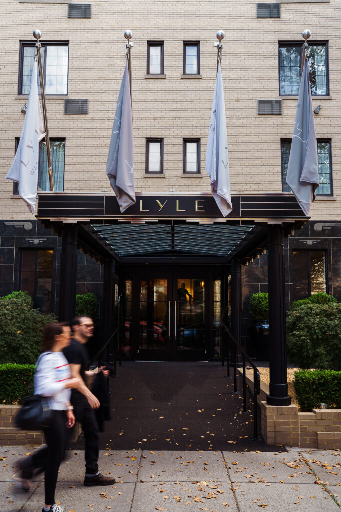 Approach to Lyle Washington DC. Five flagpoles bear Lyle branding above the entrance canopy. The canopy reads LYLE in stylised capitals on a black marble band. Twin glass doors sit beneath a glazed entry vestibule. Black marble columns flank the doorway with stylised eagle-wing relief panels at the base.