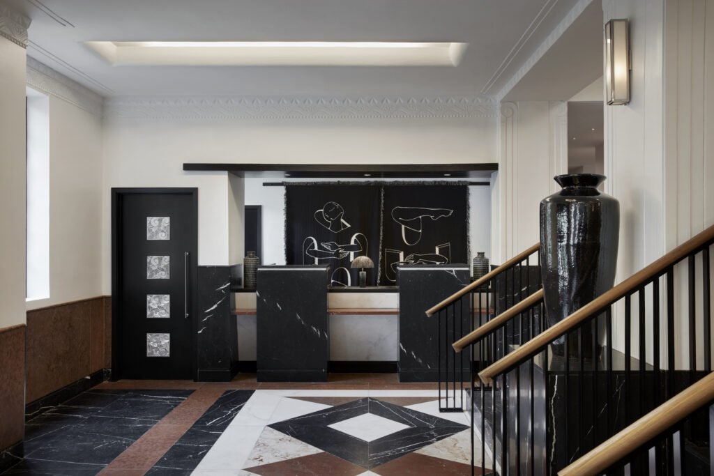 Reception area at Lyle Washington DC. Two black marble check-in podiums sit beneath a tapestry artwork depicting abstract figures in black and white. Ribbed black ceramic vases sit on a built-in shelf behind the desks. The floor is a geometric marble inlay in dark and pale tones with deeper accent stones. To the left, a black panelled door bears four art deco silver relief medallions.