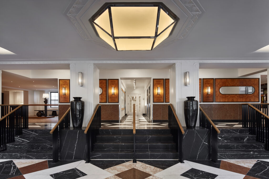 Main lobby at Lyle Washington DC. A hexagonal art deco ceiling fixture is set into a decorative plaster ceiling with frieze detail. The floor is a marble inlay in dark and pale tones with deeper accent stones. Burl-wood wall panels frame integrated sconces along both walls. Two large black ceramic urns sit atop balustrade pedestals flanking a stair down to the corridor beyond.