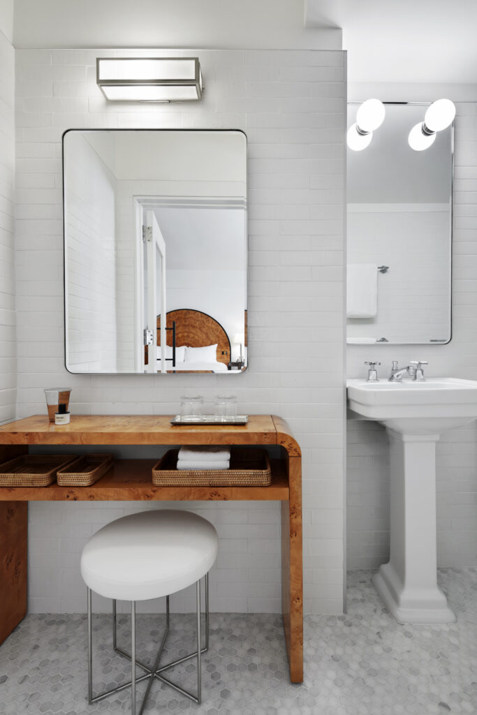 Dressing area in the Premier Corner King bathroom at Lyle Washington DC. A wall-mounted burl-wood vanity console holds woven wicker trays for amenities. Above the console, a chrome-framed rectangular mirror is set against a white-tiled wall. A round white-cushioned stool with chrome legs sits beneath the console. Hex-pattern marble mosaic floors run through the space.