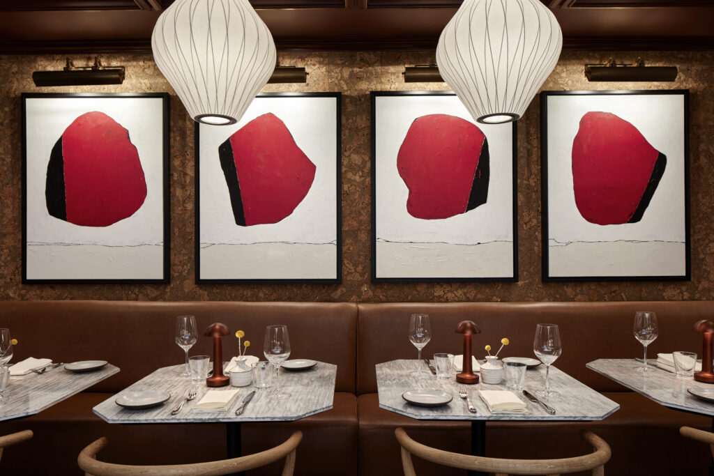 Lyle’s restaurant at Lyle Washington DC. Walnut herringbone flooring with cork-clad walls. Tan leather banquettes line the perimeter beside marble-topped tables. Wishbone chairs in natural wood sit at each table. A curved white sofa anchors the centre, with Nelson Bubble pendant lamps overhead. The open kitchen pass and backlit glassware shelving are visible to the rear.