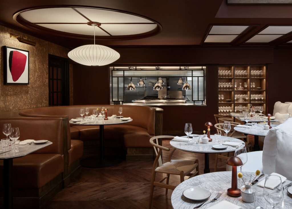 Lyle’s restaurant skylit area at Lyle Washington DC. A glass skylight runs overhead, edged with cork panelling and dark wood beams. Beneath the skylight, an oval white banquette curves around a central wooden table. Smaller marble tables with wishbone chairs surround the central seating. Multiple Nelson Bubble pendant lamps in varied shapes hang from the rafters.