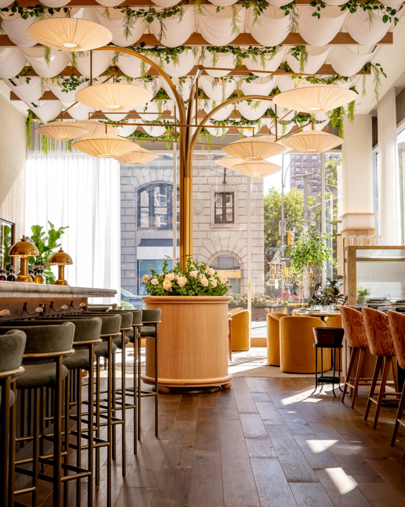 Park Rose restaurant at Hotel Park Ave. Pleated fabric canopy ceiling with hanging foliage. Brass tree-form chandelier with multiple shaded lights above a central planter of cream roses. Marble bar counter with green velvet stools on the left. Floor-to-ceiling windows onto Park Avenue. Park Rose signage visible on the front window in reverse.