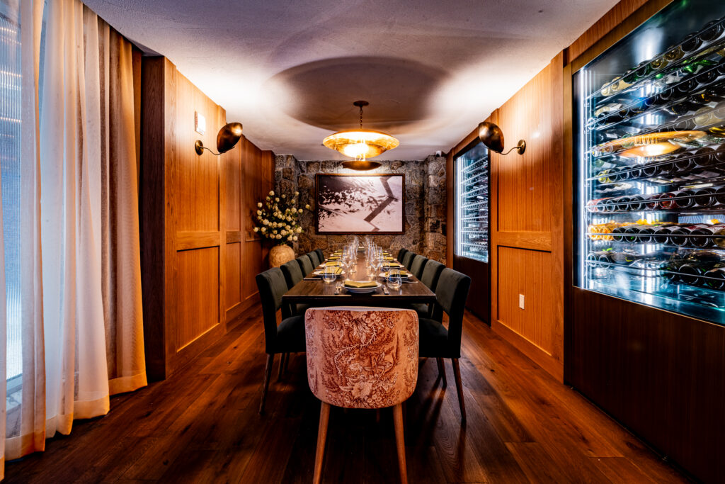 Park Rose private dining room at Hotel Park Ave. Long dark wood table set for around twelve. Wood-panelled walls with a stone-clad end wall. Backlit glass-fronted wine cabinets line the right wall. Patterned upholstered host chair at the head of the table. Brass pendant light overhead.