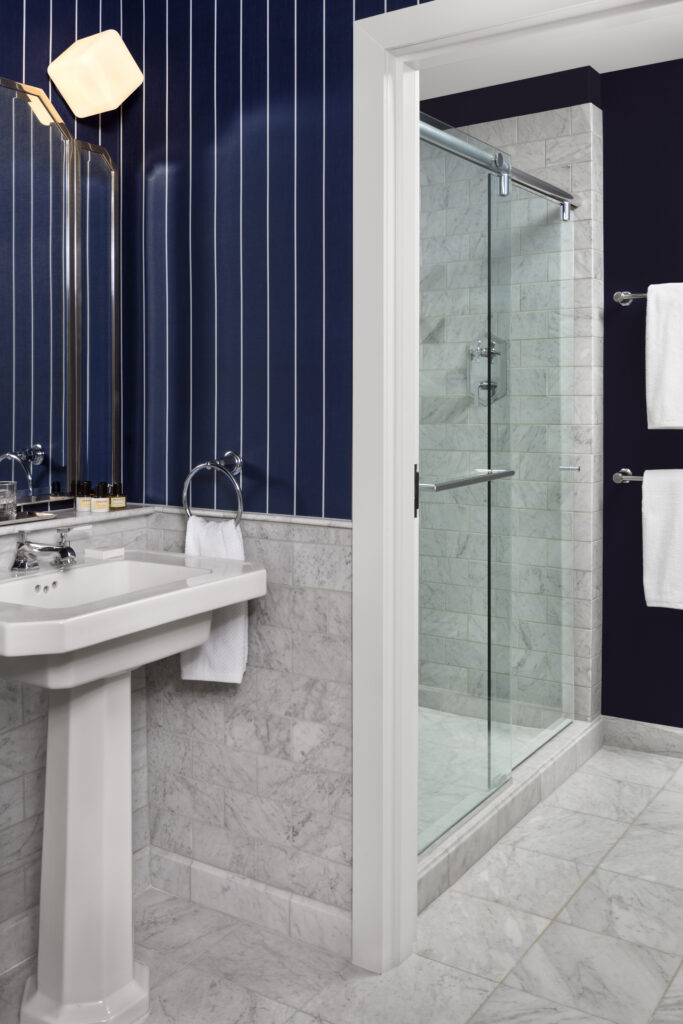Classic Room bathroom at Riggs Washington DC. White porcelain pedestal sink with chrome taps and a wall-mounted chrome towel ring. Sliding glass shower enclosure with marble subway tile and a fixed showerhead. Carrara marble floor and lower wall tiling throughout. Navy and white pinstripe wallpaper above the marble. Chrome and frosted glass wall light over the sink.