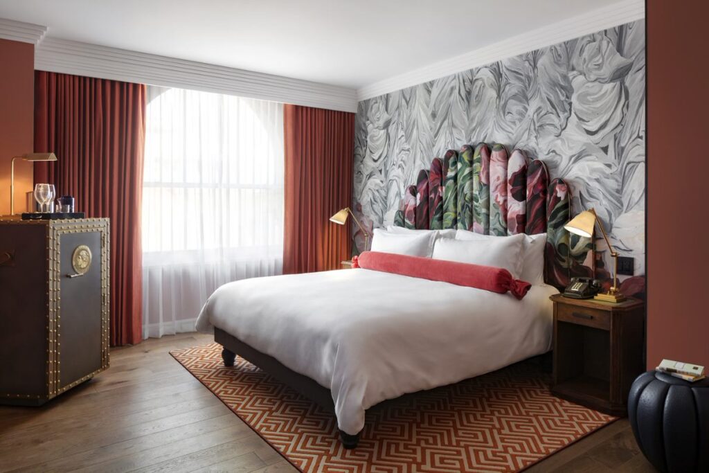 Classic Room bedroom at Riggs Washington DC. King bed with white linen and a red velvet bolster pillow at the headboard. Fluted upholstered headboard painted with a floral pattern set against marbled grey and white wallpaper. Dark wood bedside table on the right with a brass desk lamp and a corded telephone. Studded leather trunk on the left used as a minibar cabinet. Rectangular window with sheer and red velvet curtains. Patterned red and cream rug on a hardwood floor.