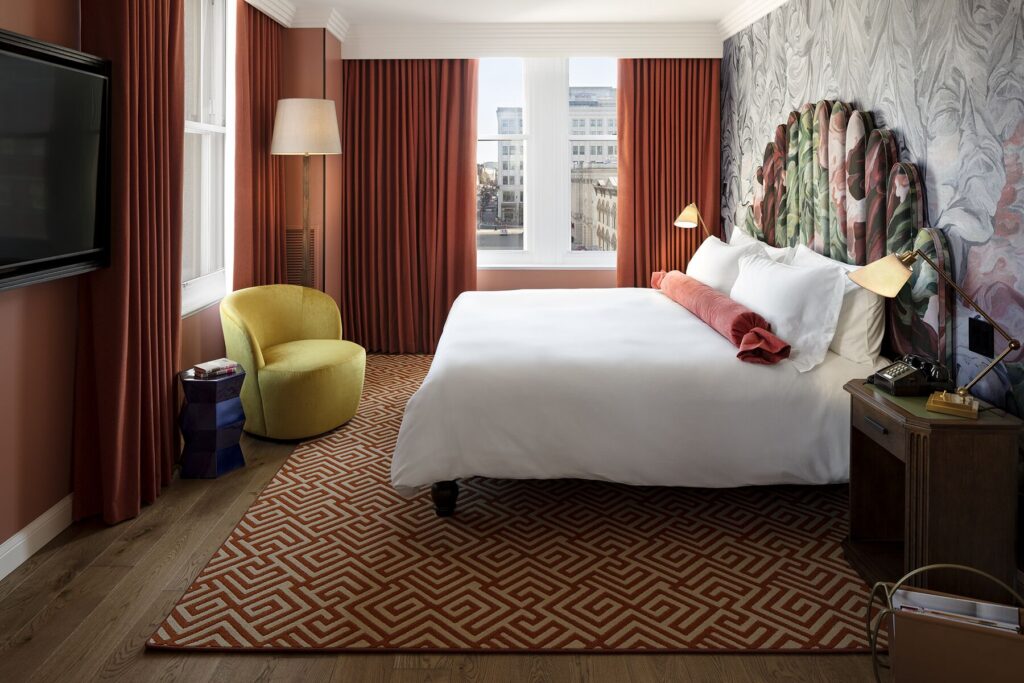 Deluxe Room bedroom at Riggs Washington DC. King bed with white linen and a coral bolster pillow facing a wall-mounted flat-screen television. Yellow velvet armchair with a brass floor lamp beside the window. Patterned red and cream rug on a hardwood floor. Bedside table on the right with a corded telephone and a brass desk lamp, with notebooks stacked beside them. Floor-to-ceiling window with sheer and red velvet curtains, with city rooftops visible outside.