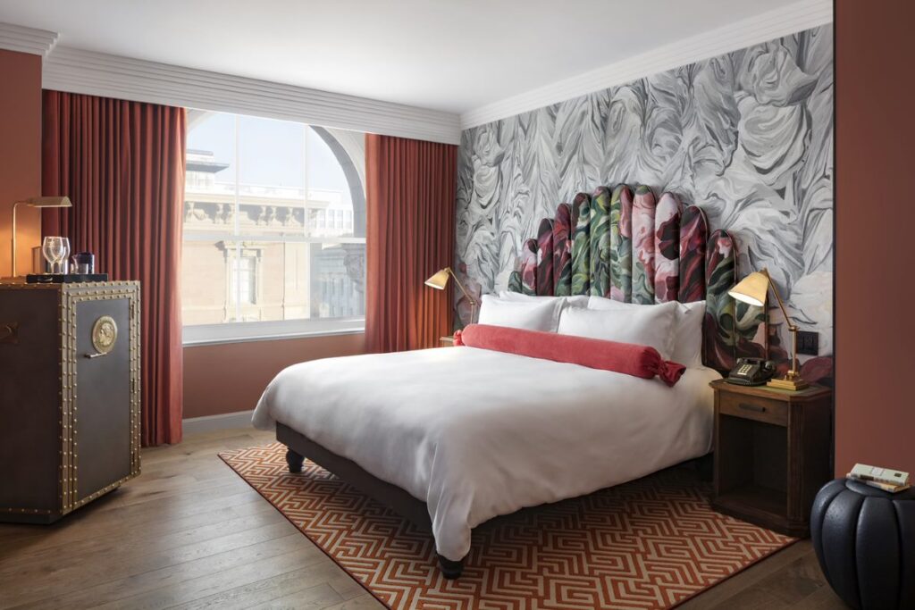 Alternate view of a Deluxe Room at Riggs Washington DC. King bed facing an arched window with sheer and red velvet curtains, with the carved stone façade of a neighbouring building visible outside. Painted floral headboard against marbled grey and white wallpaper. Studded leather trunk on the left side of the bed. Patterned red and cream rug on a hardwood floor.