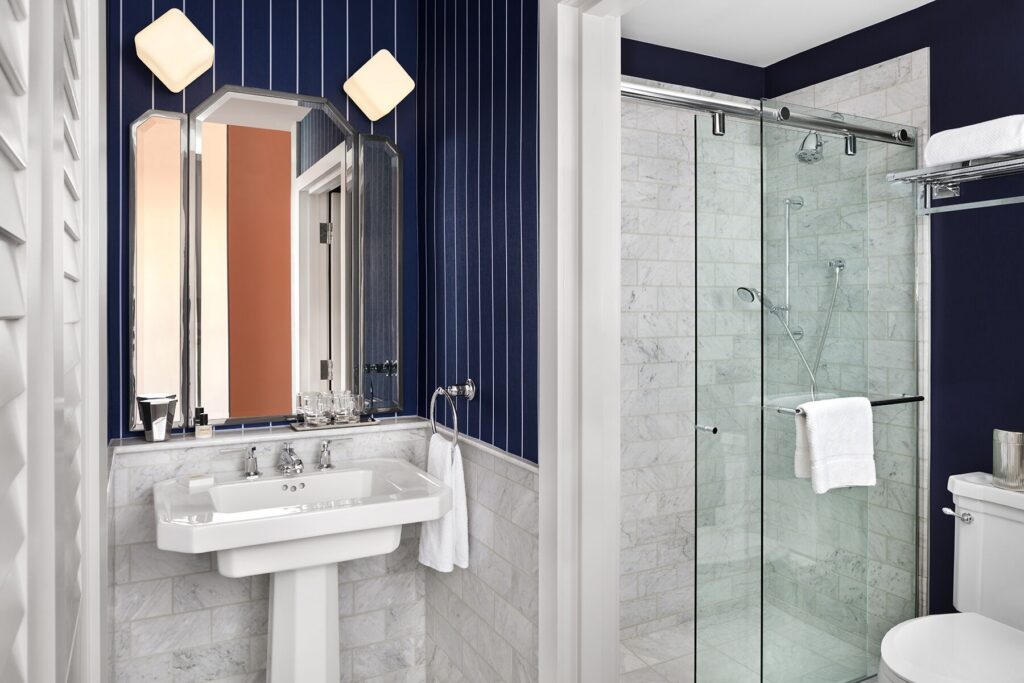 Deluxe Room bathroom at Riggs Washington DC. White porcelain pedestal sink with chrome taps below a faceted three-panel mirror in chrome frame. Two cubic frosted glass wall lights flank the mirror. Sliding glass shower enclosure with marble subway tile, fitted with both a fixed showerhead and a handheld showerhead on a slide rail. Toilet visible to the right of the shower. Carrara marble floor and lower walls. Navy and white pinstripe wallpaper above the marble.