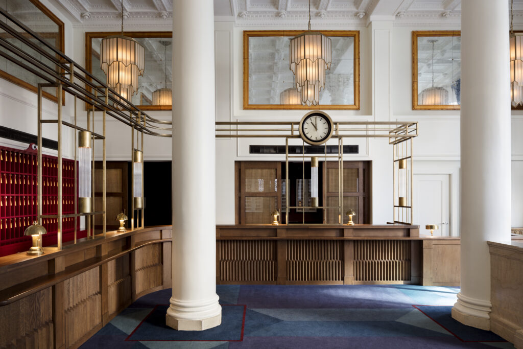Reception area at Riggs Washington DC. Curved oak reception desk with two table lamps and a brass and steel structure overhead supporting an analogue wall clock. Red key wall visible on the left with brass key cabinets. Green marble Corinthian columns flank the space under a coffered white ceiling. Geometric blue and red carpet on the floor.