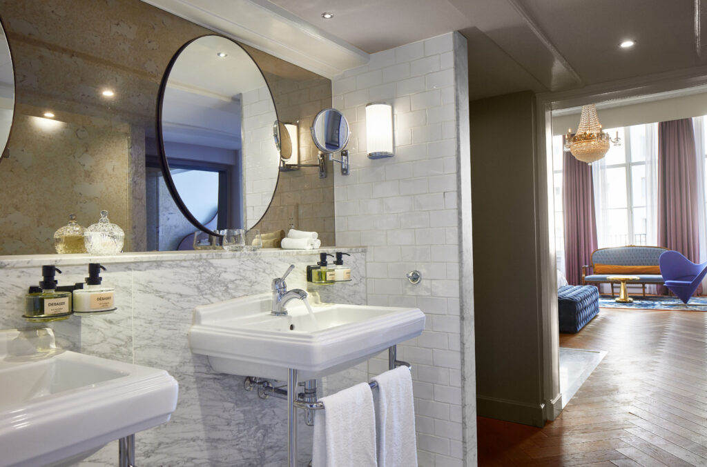 En-suite bathroom at Pulitzer Amsterdam. Wall-mounted ceramic basin with single chrome tap on marble splashback. Round black-framed mirror above. Magnifying mirror wall-mounted to the right. Wall-mounted DEBASER amenity dispensers (D.S. & Durga). White subway tile walls. Adjoining bedroom visible through doorway at right.