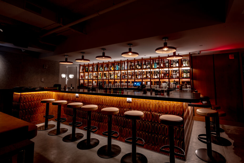 Seed Library bar at Hotel Park Ave. Long counter with a patterned glazed tile front in amber. Backbar shelving uplit from beneath holds the spirits range. Six pendant lamps over the bar. Upholstered round-topped stools at the counter. Exposed ceiling with track lighting.