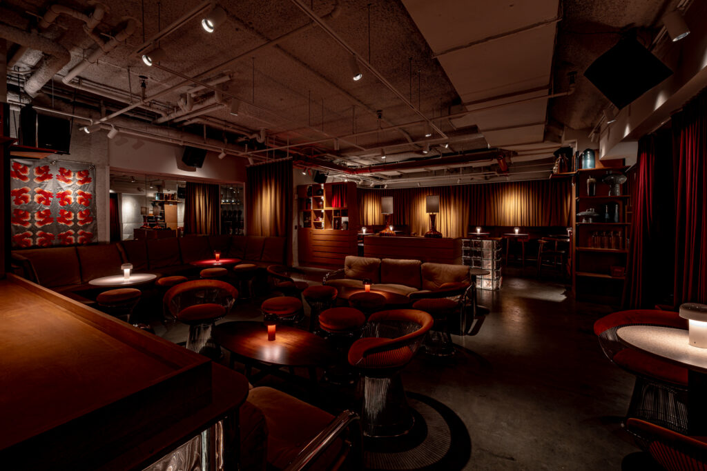 Seed Library lounge at Hotel Park Ave. Curved red velvet banquette seating with cushions wraps the back wall. Wooden chairs at low tables with rechargeable table lamps. Bar visible at the rear under pendant lighting. Curtains along the back wall. Exposed concrete ceiling with track lighting.