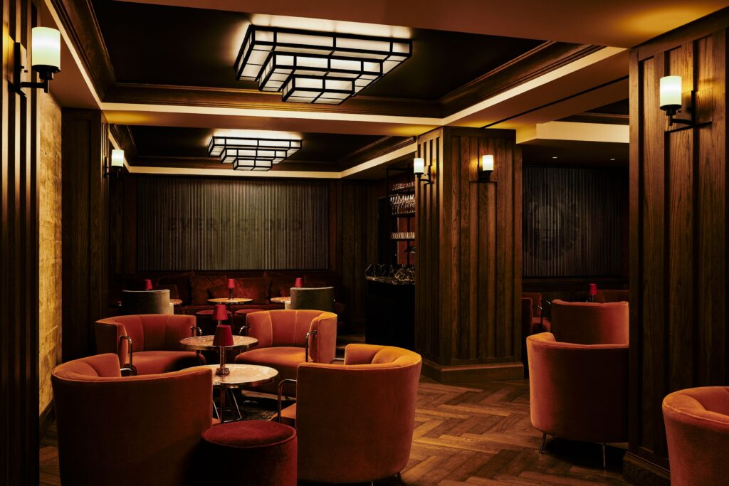Lounge seating area at Silver Lyan. Orange velvet tub chairs arranged around small round tables with red shaded table lamps. Herringbone wood floor. Wood panelling and a low banquette with cushions along the back wall. Geometric ceiling fittings in black metal provide the main lighting. Brass and frosted glass wall sconces.