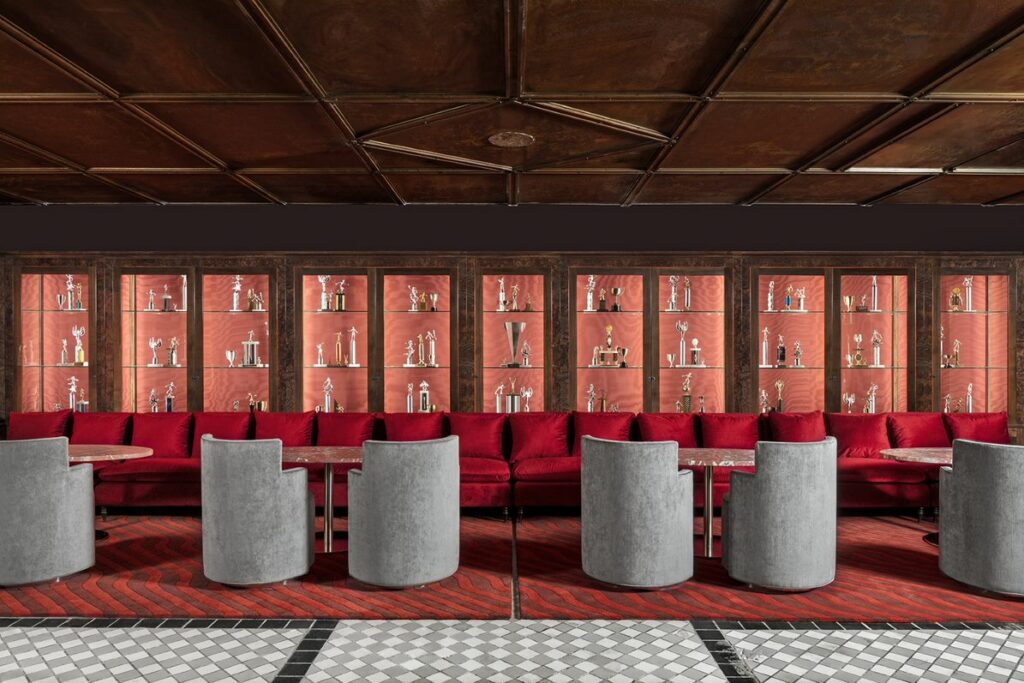 Trophy wall feature at Silver Lyan. Continuous red velvet banquette set in front of illuminated burr wood cabinets displaying silver and gold trophies on coral pink backing. Grey velvet tub chairs paired with round marble tables face the banquette. Red and pink chevron carpet meets a black and white checkerboard floor. Wood-panelled ceiling above.