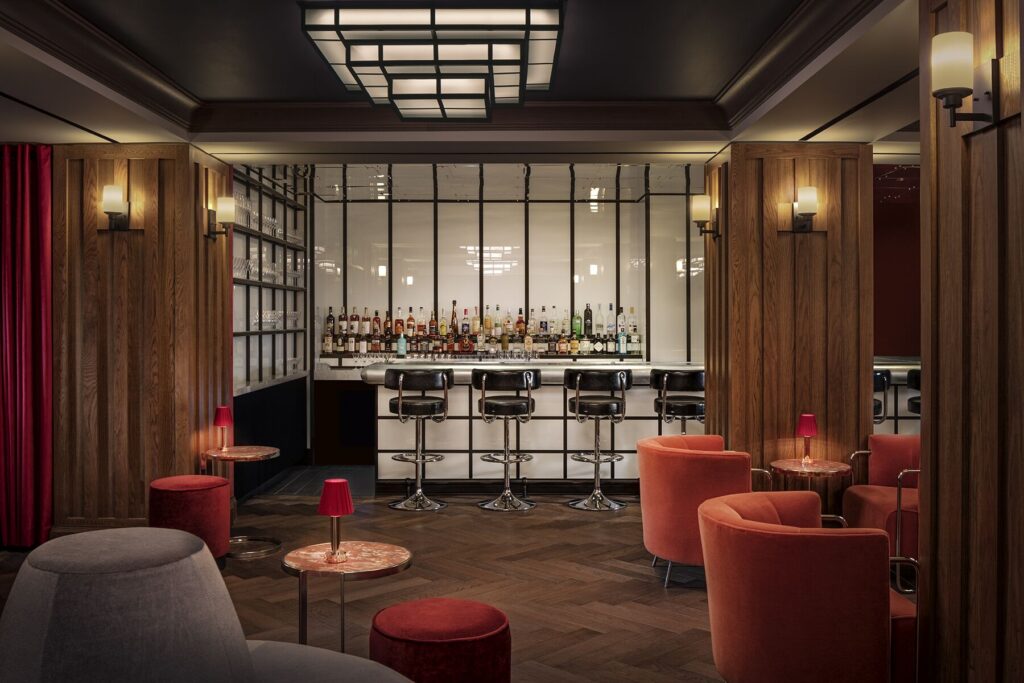 The bar at Silver Lyan, the basement cocktail bar at Riggs Washington DC by Ryan Chetiyawardana. Caged metal bar back holds bottles behind a white marble counter. Four black leather bar stools at the bar. Wood panelling and red velvet curtains frame the space. Geometric ceiling pendant in black metal. Herringbone wood floor.
